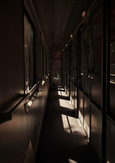 A photo looking along the interior of a train corridor. Windows on the left let in sunllight, creating deep shadows. Closed compartment doors with small windows line the right side of the corridor.