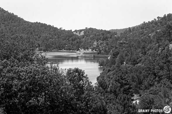 A serene black-and-white landscape view showcasing a tranquil reservoir surrounded by lush green hills and trees, with a wooden pier stretching out into the water. The hills rise gently, creating a peaceful, natural atmosphere.