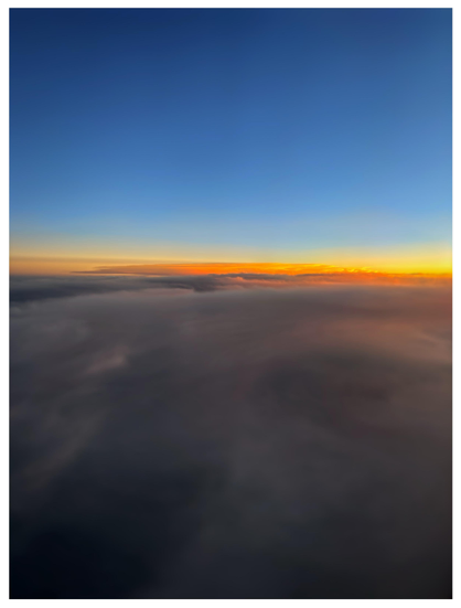 Photo taken from an airplane window at sunset. A field of fog-like gray clouds tinged with gold stretches to the horizon: a luminous orange band that splits the image in half. The sky above the clouds is a clear, deepening blue.