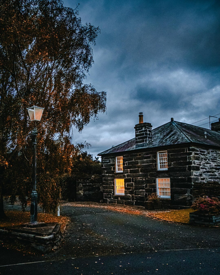 A traditional Welsh stone cottage with four white sash windows at twilight. The bottom left window is lit up from inside casting a bright orange glow. To the left, an ornate, black iron street lamp stands at the edge of a curved driveway in front of a tall tree with autumnal orange foliage. Overhead the twilight sky is dark and overcast with grey and blue clouds.