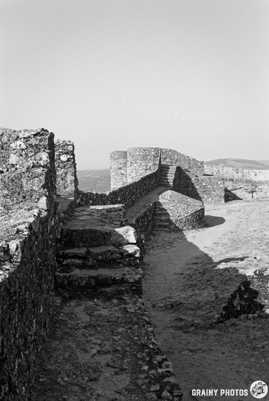 A black-and-white photograph depicts a stone staircase leading upwards within the walls of an old fortress. The steps are worn and uneven, with a long shadow stretching across the ground, suggesting morning light. The scene evokes a sense of history and quiet solitude.