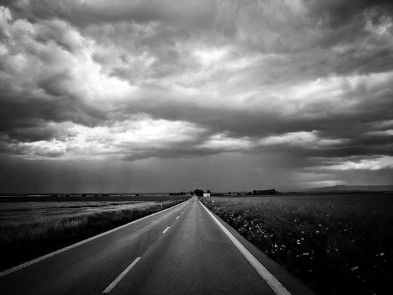 A black-and-white photograph of a straight, empty two-lane road extending into the distance, viewed from road level along its centre line. Flat grassy fields border the road on both sides. The sky is covered with thick, heavy clouds in dark tones, creating strong contrast with the lighter road surface. A few structures and sparse trees are visible on the horizon. The perspective converges at a vanishing point, with the road edges narrowing towards it.