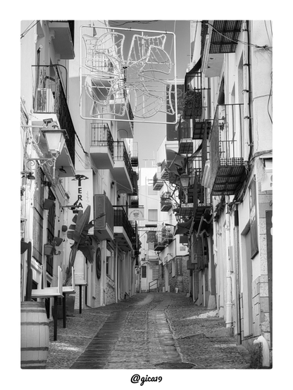 Esta foto la hice por la mañana del pasado 3 de septiembre en la zona antigua de Peñíscola, en la provincia de Castellón. Es una imagen en blanco y negro, en formato vertical, de una calle estrecha y empedrada que sube suavemente entre casas encaladas.
El suelo es de adoquines irregulares y tiene en el centro una franja de piedras más alargadas que marca el trazado de la calle. Su superficie se ve ligeramente húmeda y brillante. Al fondo, la calle gira y sigue hacia arriba entre más edificios blancos. En primer plano, abajo a la derecha, hay una tapa de alcantarilla circular. En la esquina inferior izquierda se ve un barril de madera.
A ambos lados se alinean fachadas blancas con muchos balcones de forja. Algunos tienen suelos de azulejos con dibujos de cuadros y otros llevan toldos o plantas. Hay farolas de hierro en las paredes, tuberías blancas que bajan por las fachadas y cables que cruzan de un lado a otro. En la parte izquierda hay rejas en las ventanas, un aparato de aire acondicionado en un balcón y una gran hoja de platanera que sobresale junto a la pared.
Sobre la calle cuelga una gran estructura rectangular de luces decorativas con forma de flor, y en el centro se ve un cielo claro. Entre los carteles de los comercios se leen «Tattoo», «La Casa de las Toallas», «Galatea» y un cartel de un «Magic Museum» con el rostro de una persona. A la izquierda hay mesas y sillas apiladas junto a los locales, y la calle está sin gente. La luz de la mañana crea zonas de sombra y de claridad sobre las paredes.