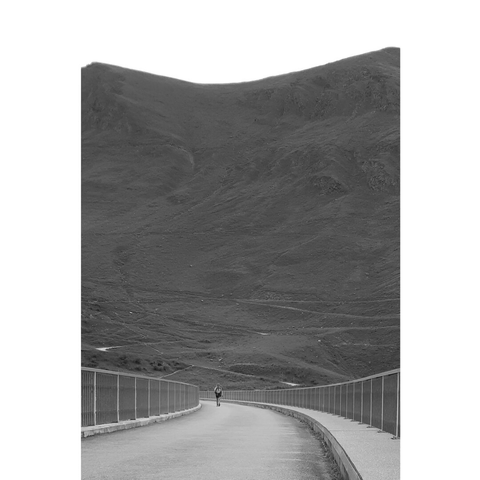 Black and white picture of a tiny lonely person walking on the curved road above the dam forming the lake Moiry. Gray mountains are in the background.