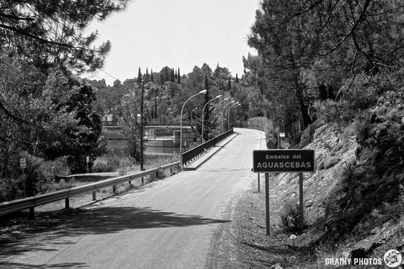 A black and white image of a winding road across the reservoir dam, featuring a sign for "Embalse del Aguascebas." The landscape is surrounded by trees, creating a serene, natural setting.