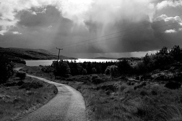 Eine schmale Straße führt in einer kleinen Kurve auf einen See zu. Am Himmel sind ordentlich Wolken, aus den der Regen fällt, aber auch viel Licht, das die Wasseroberfläche zum Funkeln bringt. Im Vordergrund rechts und links Gräser, Bäume und Büsche, im Hintergrund Hügel und Berge.
A narrow road leads to a lake in a small curve. There are a lot of clouds in the sky, from which the rain falls, but also a lot of light, which makes the water surface sparkle. In the foreground right and left grasses, trees and bushes, in the background hills and mountains.