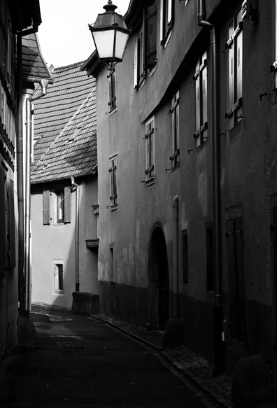 A black and white image shows a narrow cobblestone street with old buildings on either side. An old lantern hangs over the street.