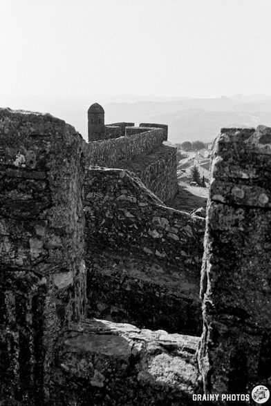 A black-and-white photograph shows a stone fortress viewed through an arched opening, with rolling hills visible in the distance. The light suggests morning, illuminating the textured stone walls and creating a sense of history and solitude.
