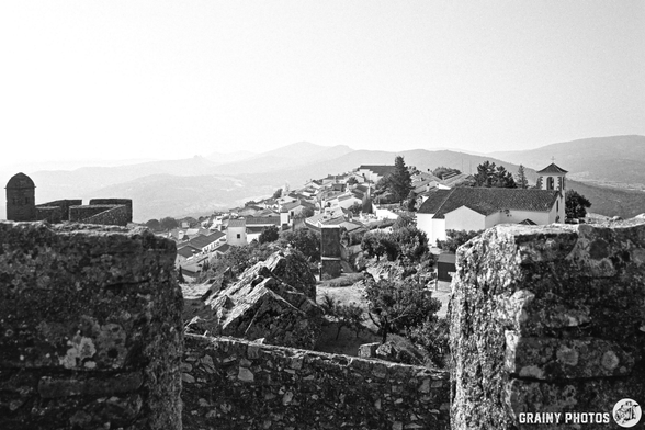 A black-and-white photograph shows a traditional Portuguese village, Marvão, situated amongst rolling hills. Stone walls frame the view, leading to a cluster of white buildings with red-tiled roofs and a church steeple in the distance, all bathed in morning light.