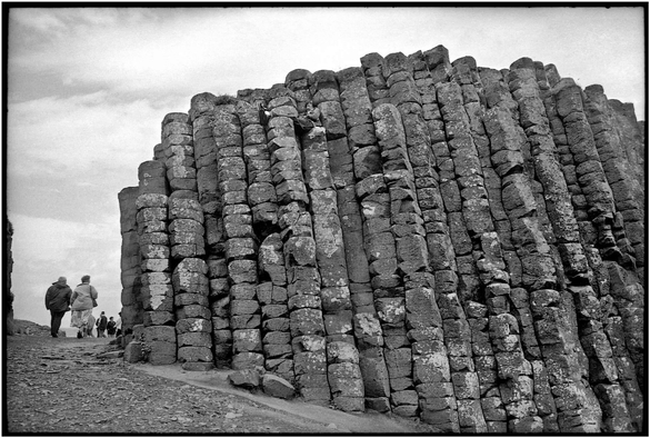 En n&b, sur le site de la Chaussée des Géants en Irlande du Nord, un mur de colonnes basaltiques longé à gauche par un sentier sur lequel passent quelques touristes. Un personnage allongé est camouflé sur le haut du mur. / A black-and-white photograph taken at the Giant’s Causeway in Northern Ireland, showing a wall of basalt columns with a path running alongside it on the left, along which a few tourists are walking. A figure lying down is hidden at the top of the wall.
