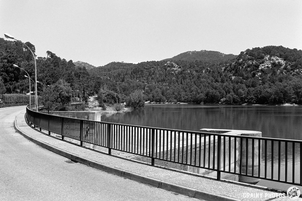A curved road runs alongside a tranquil Embalse del Aguascebas surrounded by lush green hills. A black railing borders the road, reflecting the calm waters. The scene is captured in monochrome, showcasing the serene landscape and nature's beauty.