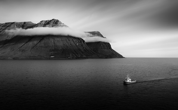 This is a monochrome image of a boat, mountains and clouds in Isafjordur, in Iceland. The thin ribbon of clouds wrap themselves around the top of the mountains, giving them a feeling of being some kind of headband. The small boat, heading towards the viewer, pulls the eye down to the corner of the image.