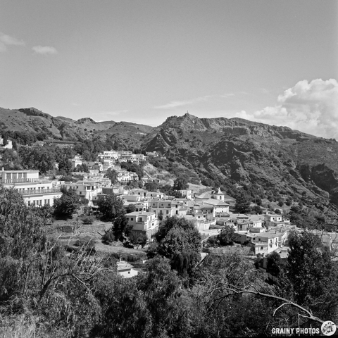 A picturesque village is situated within a valley of gently sloping, tree-covered hills. The scene is bathed in bright afternoon light, creating a tranquil and expansive view. A few buildings with white walls and terracotta roofs are visible amongst the greenery.