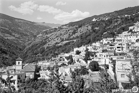 A black and white photograph captures a picturesque Pampaneira clinging to the steep slopes of a valley. Traditional white buildings with red-tiled roofs cascade down the hillside, framed by lush greenery and a partly cloudy sky. The villages of Bubion and Capileira are visible in the distance. The scene evokes a sense of tranquillity and timeless beauty, with the afternoon light softly illuminating the landscape.