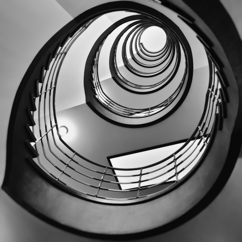 Schwarzweiss-Foto eines Treppenhauses in einem älteren Haus in Hamburg. Vom Erdgeschoß nach oben gesehen. Der Handlauf ist aus Metall.